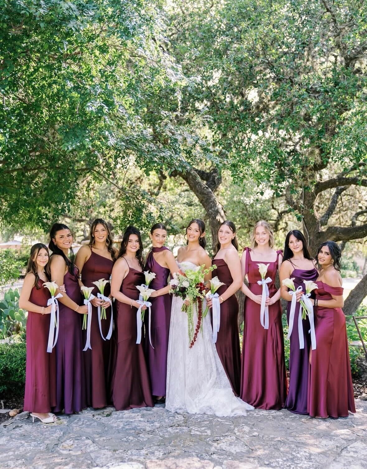 Bridesmaid party in burgundy and wine satin gowns under a live oak, each holding a single-stem calla bouquet