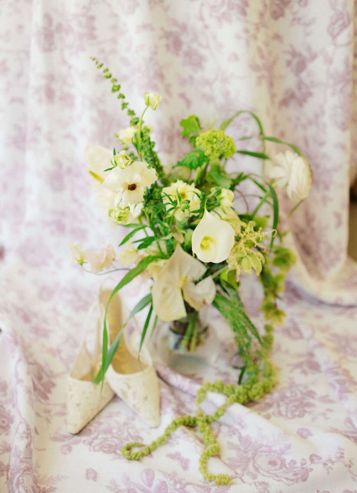 Editorial ivory and green bouquet with calla lilies, ranunculus, and trailing amaranthus next to bridal heels