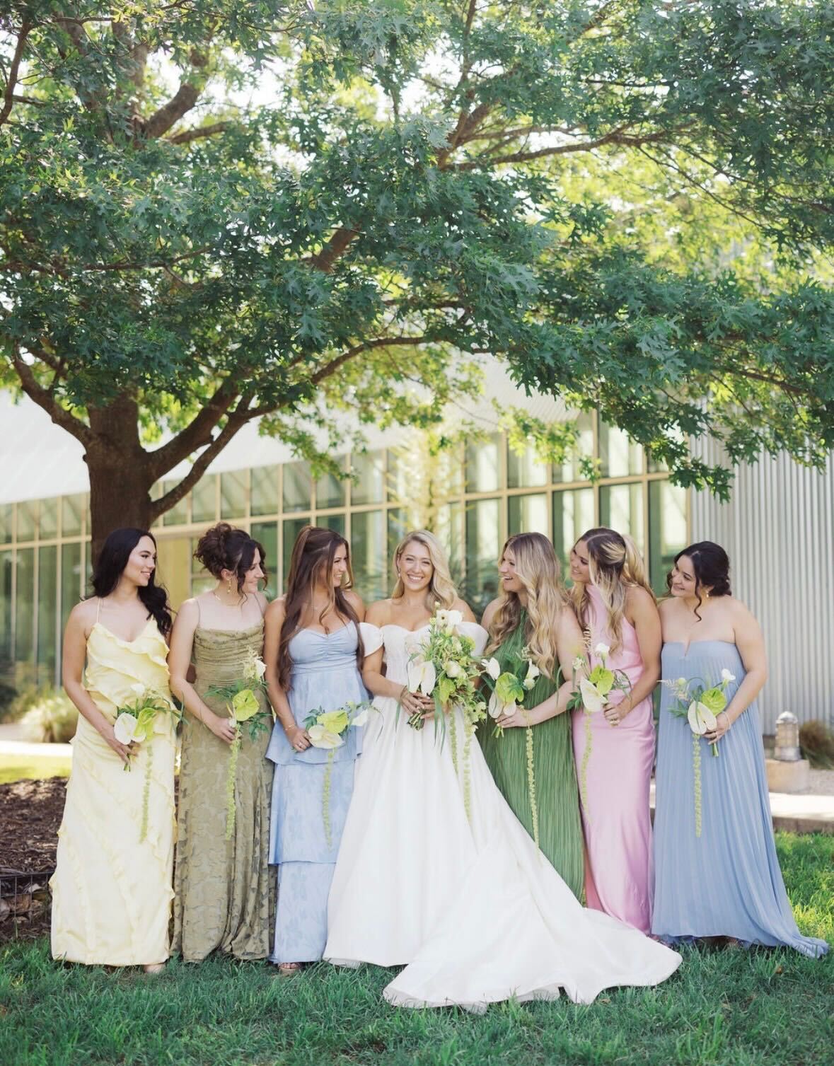Bridal party in pastel gowns each holding a small white-and-green bouquet
